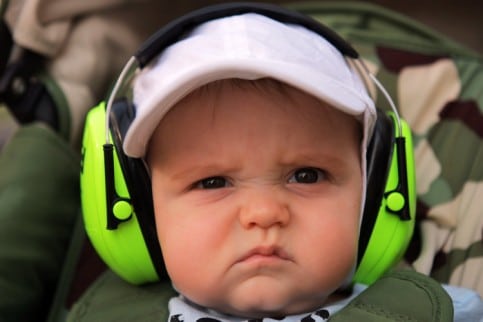 Ein Baby mit weißer Mütze und hellgrünen Kopfhörern mit Geräuschunterdrückung runzelt die Stirn und scheint verärgert oder unzufrieden zu sein.