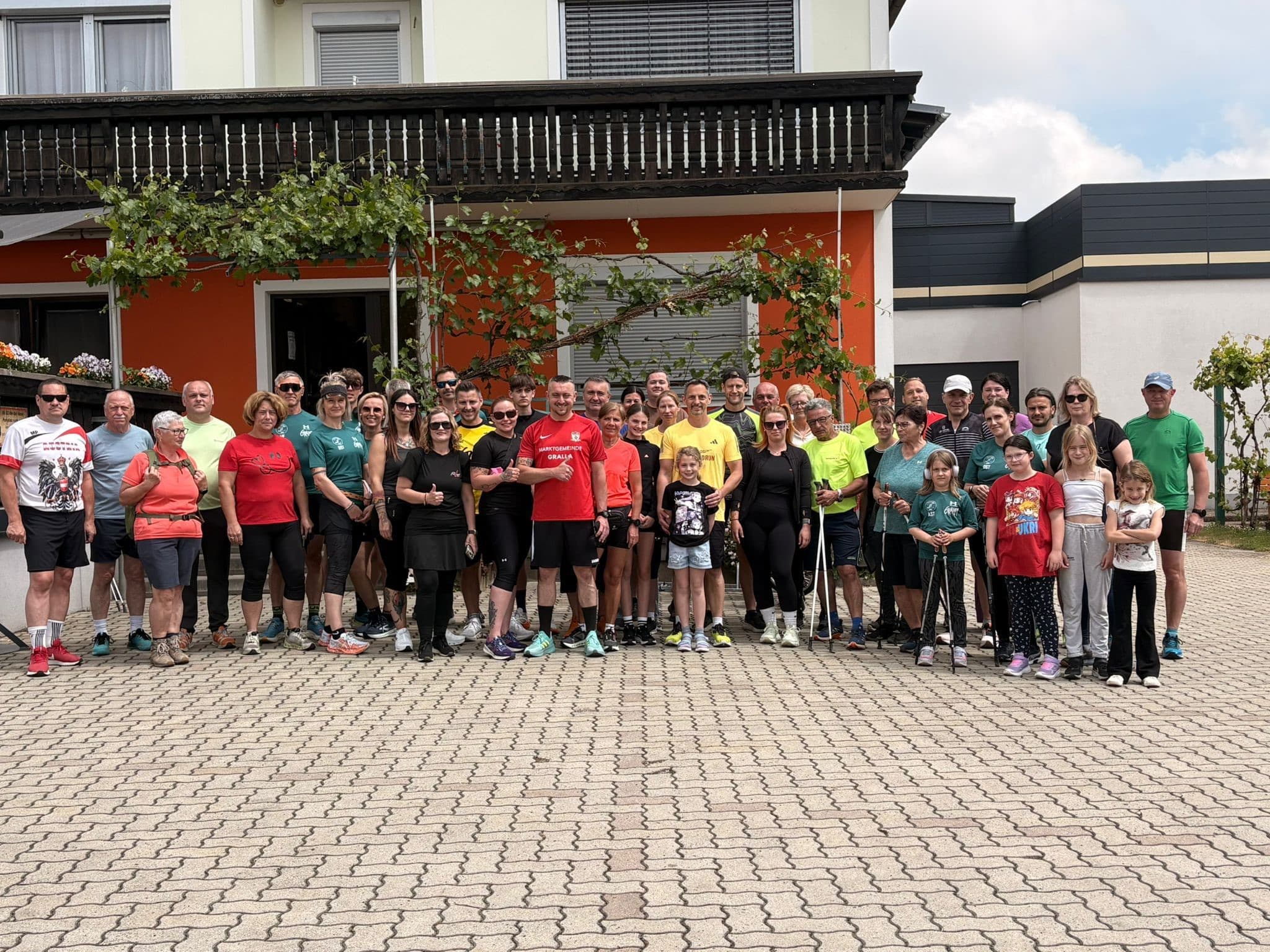 Eine große Gruppe von Erwachsenen und Kindern in Sportkleidung steht an einem sonnigen Tag lächelnd vor einem bunten Gebäude.