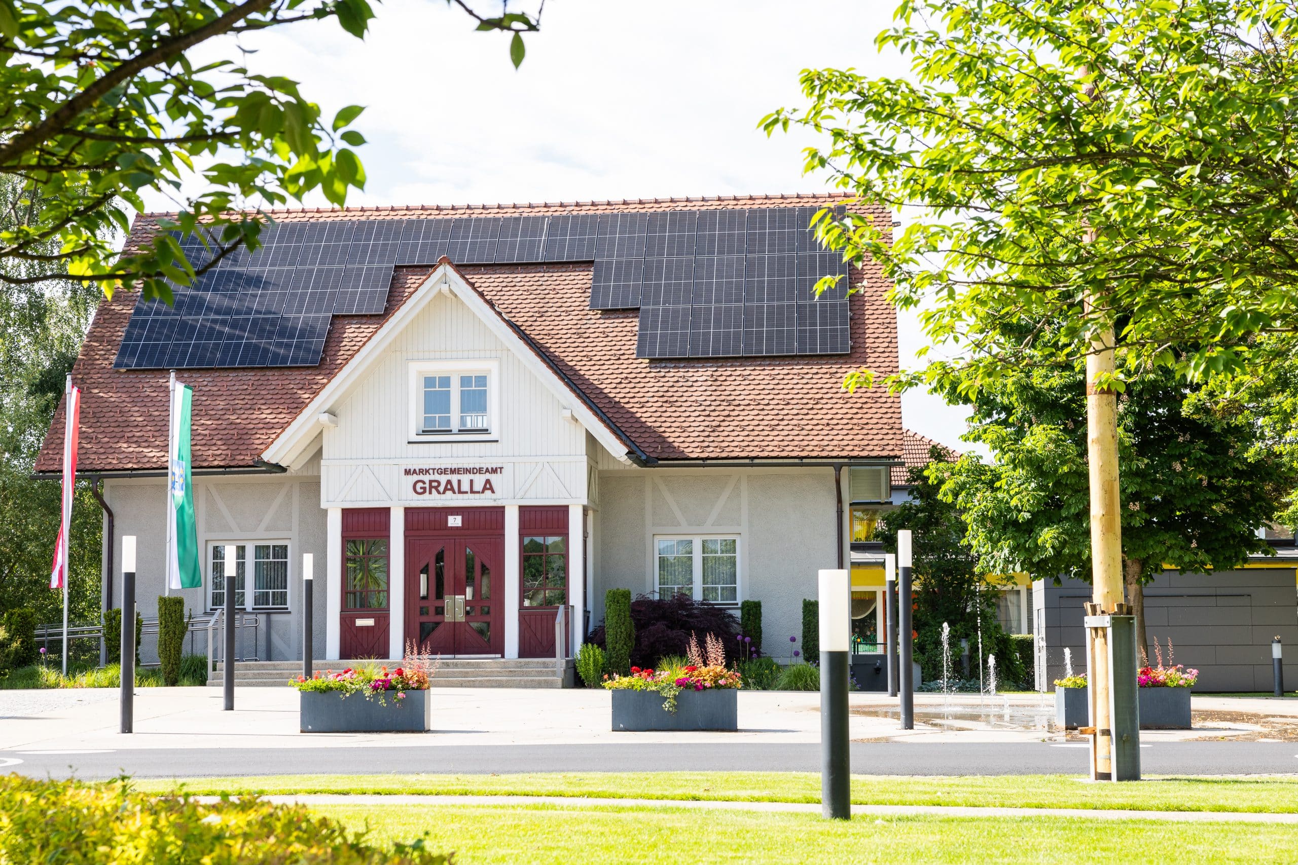 Ein Gebäude mit Solarzellen auf dem Dach, das mit "GRALLA" beschriftet ist, umgeben von Bäumen, Pflanzkübeln und Fahnen an einem sonnigen Tag.