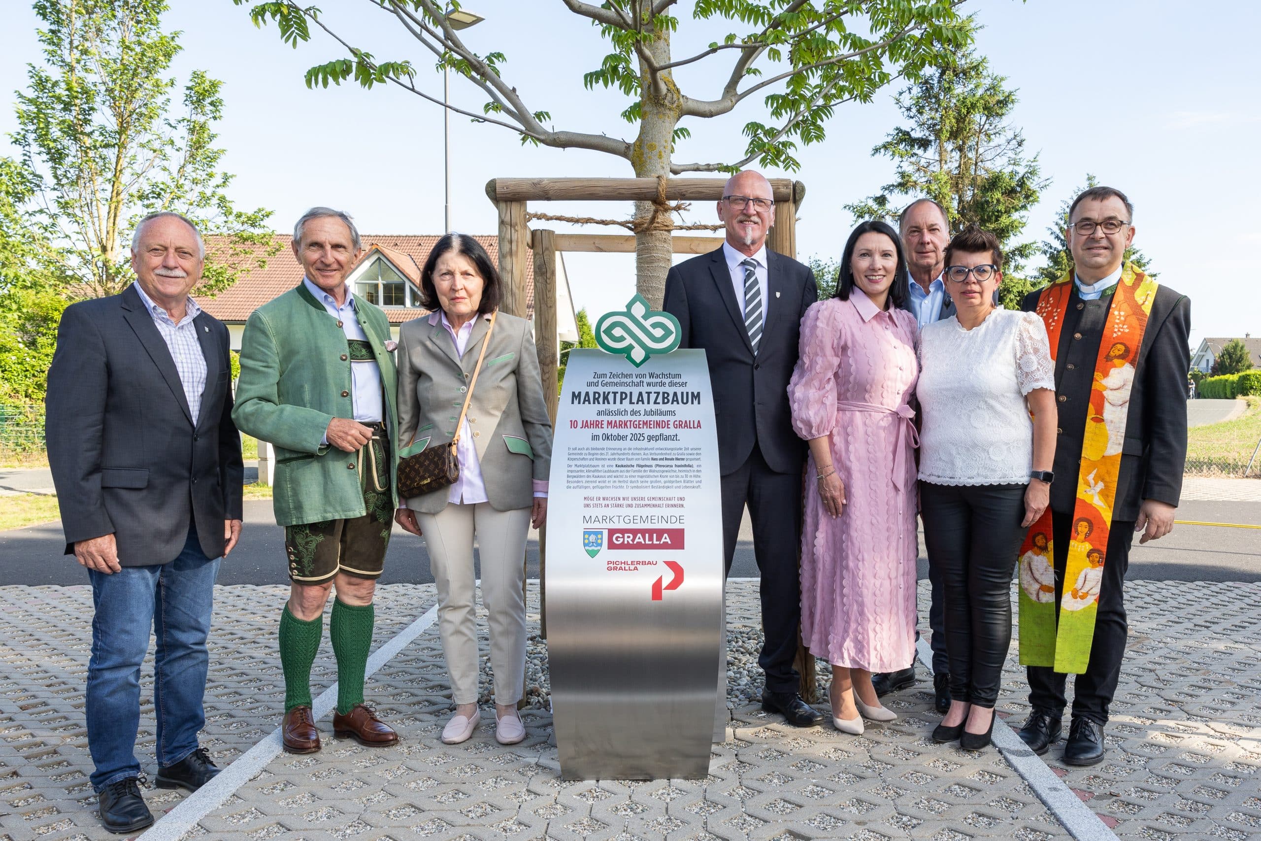 Eine Gruppe von acht Personen steht im Freien um einen Baum und ein Schild mit der Aufschrift "Marktplatzbaum", alle lächeln und sind formell und traditionell gekleidet.