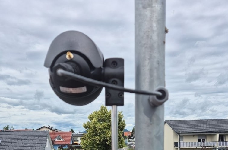 Eine schwarze Überwachungskamera, die an einem Metallmast befestigt ist und auf ein Vorstadtviertel mit Häusern und Bäumen unter einem bewölkten Himmel blickt.