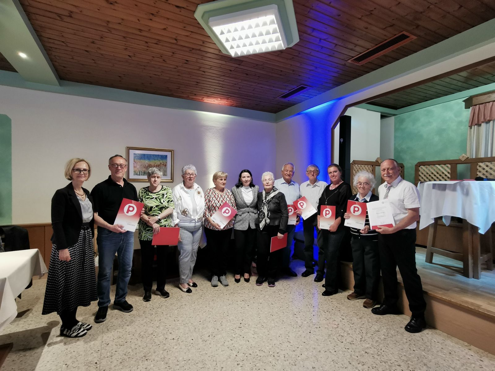 Eine Gruppe von dreizehn Personen steht in einem Raum, einige halten Urkunden und rote Mappen in der Hand und posieren und lächeln für ein Gruppenfoto.