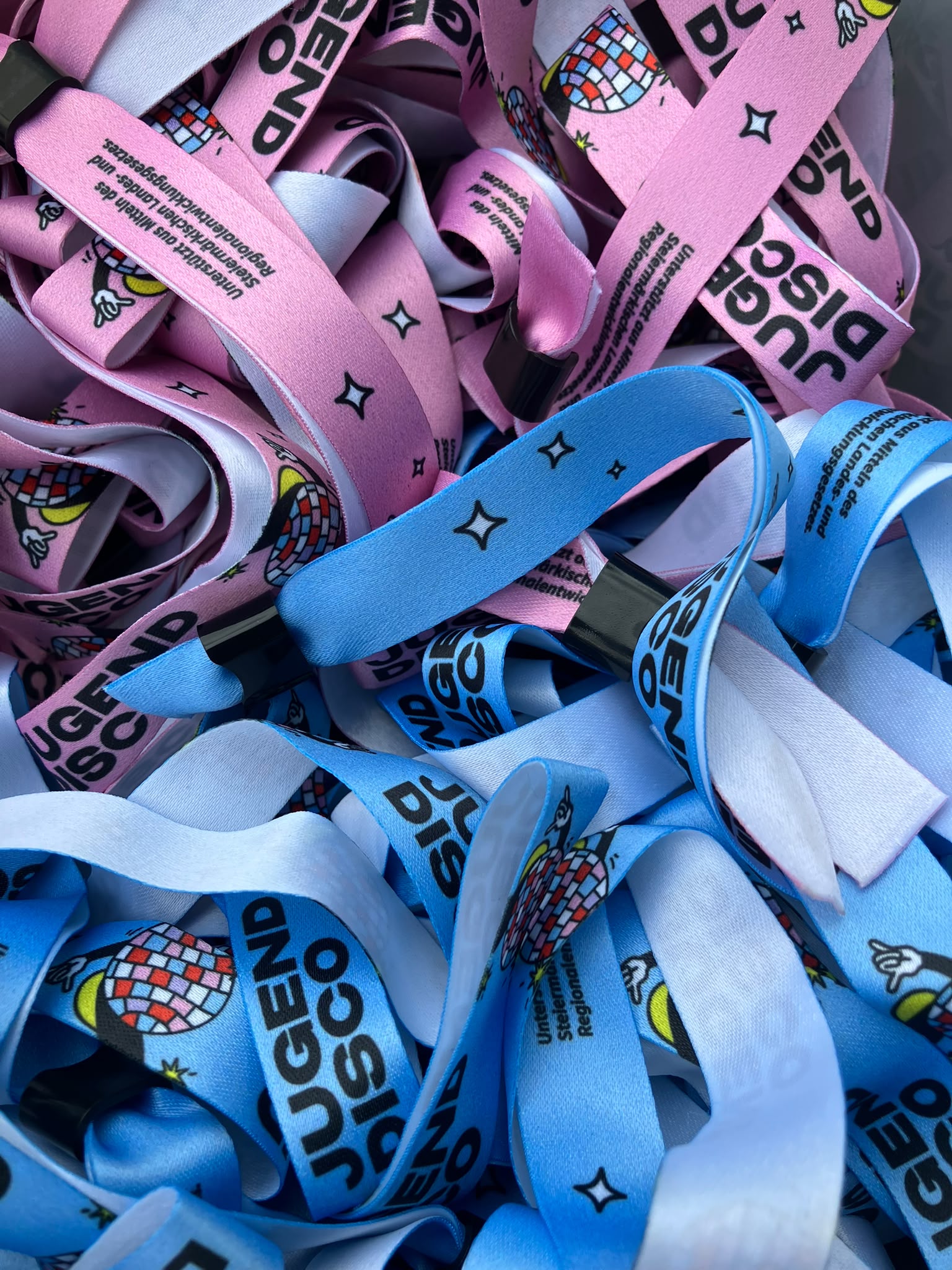 Rosa und blaue Armbänder mit der Aufschrift „JUGEND DISCO“, einer Discokugel und Sternen liegen übereinander gestapelt. Auf einigen sind neben der Discokugel Engelsflügel abgebildet.
