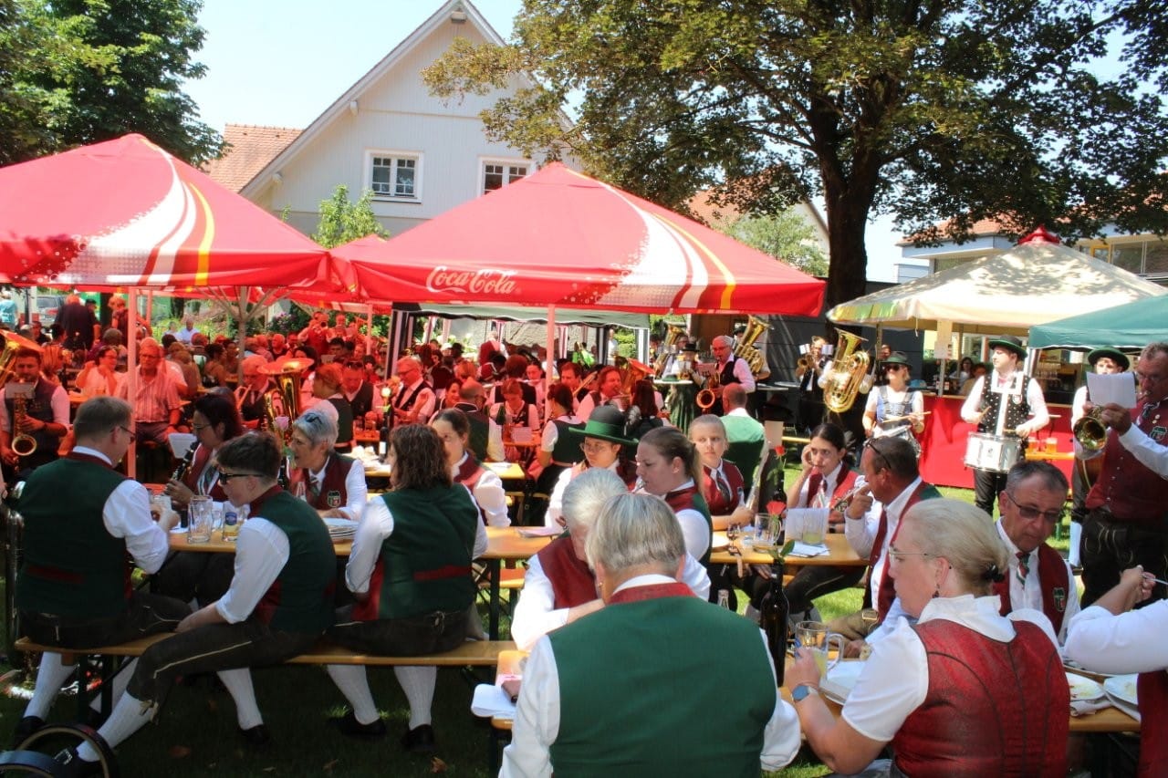 Menschen in traditioneller bayerischer Tracht sitzen unter Sonnenschirmen an Picknicktischen im Freien und genießen Speisen und Getränke, während eine Blaskapelle spielt.