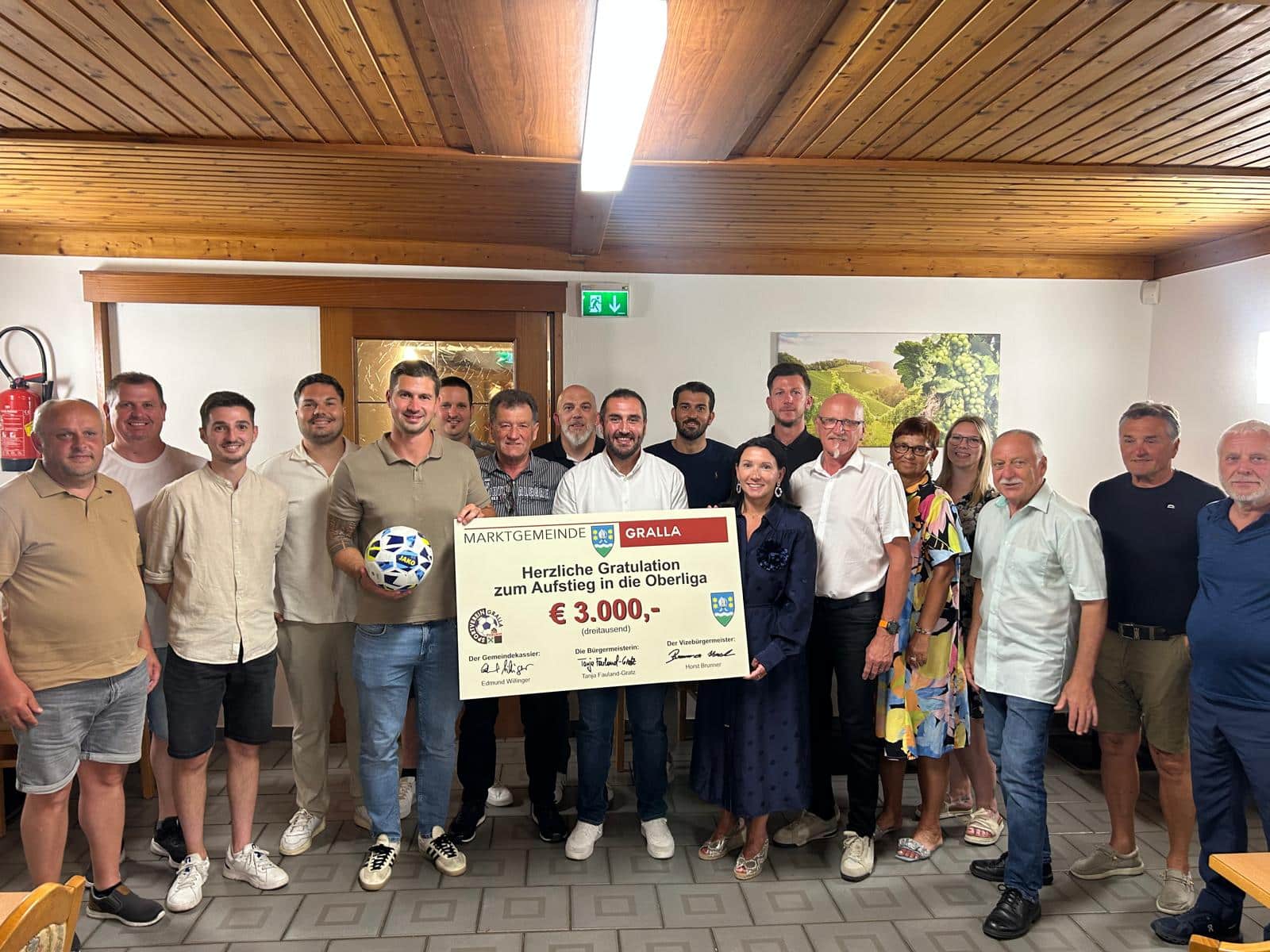 Eine Gruppe von Menschen in einem Raum hält einen großen Scheck über 3.000 Euro und einen Fußball in den Händen, lächelt und posiert für ein Foto zur Feier des Anlasses.