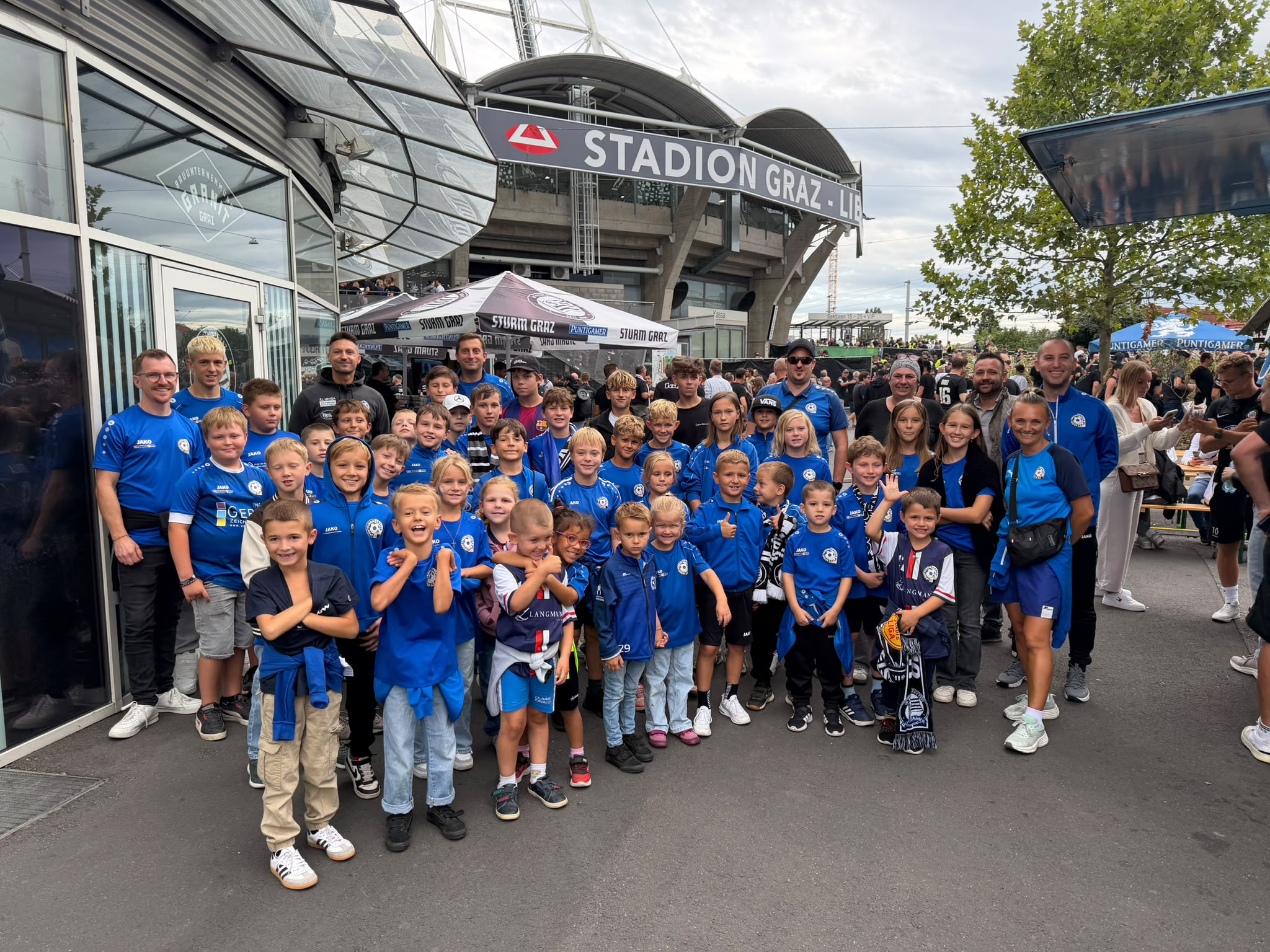 Eine große Gruppe von Kindern und Erwachsenen in blauen T-Shirts posiert lächelnd vor dem Stadion Graz, im Hintergrund sind das Stadion und die Zuschauermenge zu sehen.