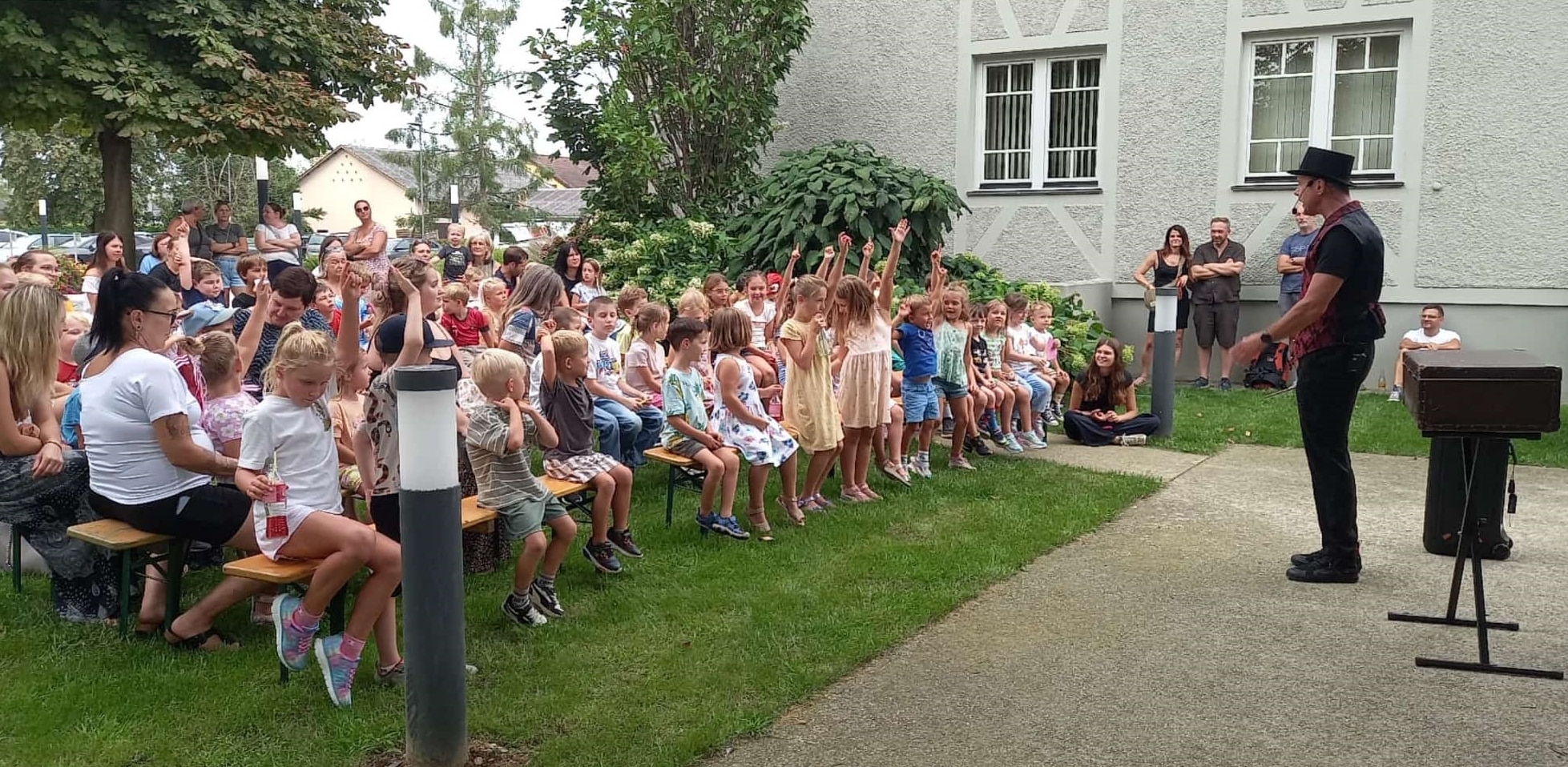 Ein Mann mit einem schwarzen Hut tritt vor einer Gruppe von Kindern auf, die draußen auf Bänken sitzen; einige Kinder heben eifrig die Hände.