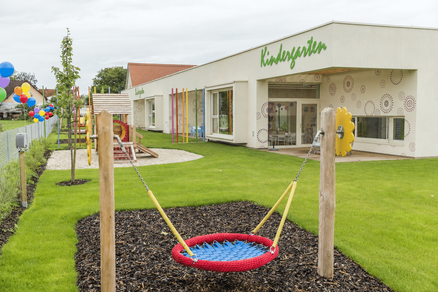 Dies ist ein moderner Kindergarten mit einem Spielplatz, einer Rasenfläche, Schaukeln, bunten Luftballons und einem einladenden Eingangsbereich, in dem kleine Kinder lernen und spielen können.