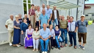 Eine Gruppe Erwachsener posiert gemeinsam im Freien vor einem Gebäude; einige stehen, andere sitzen, und ganz vorne in der Mitte sitzt ein Mann im Rollstuhl.