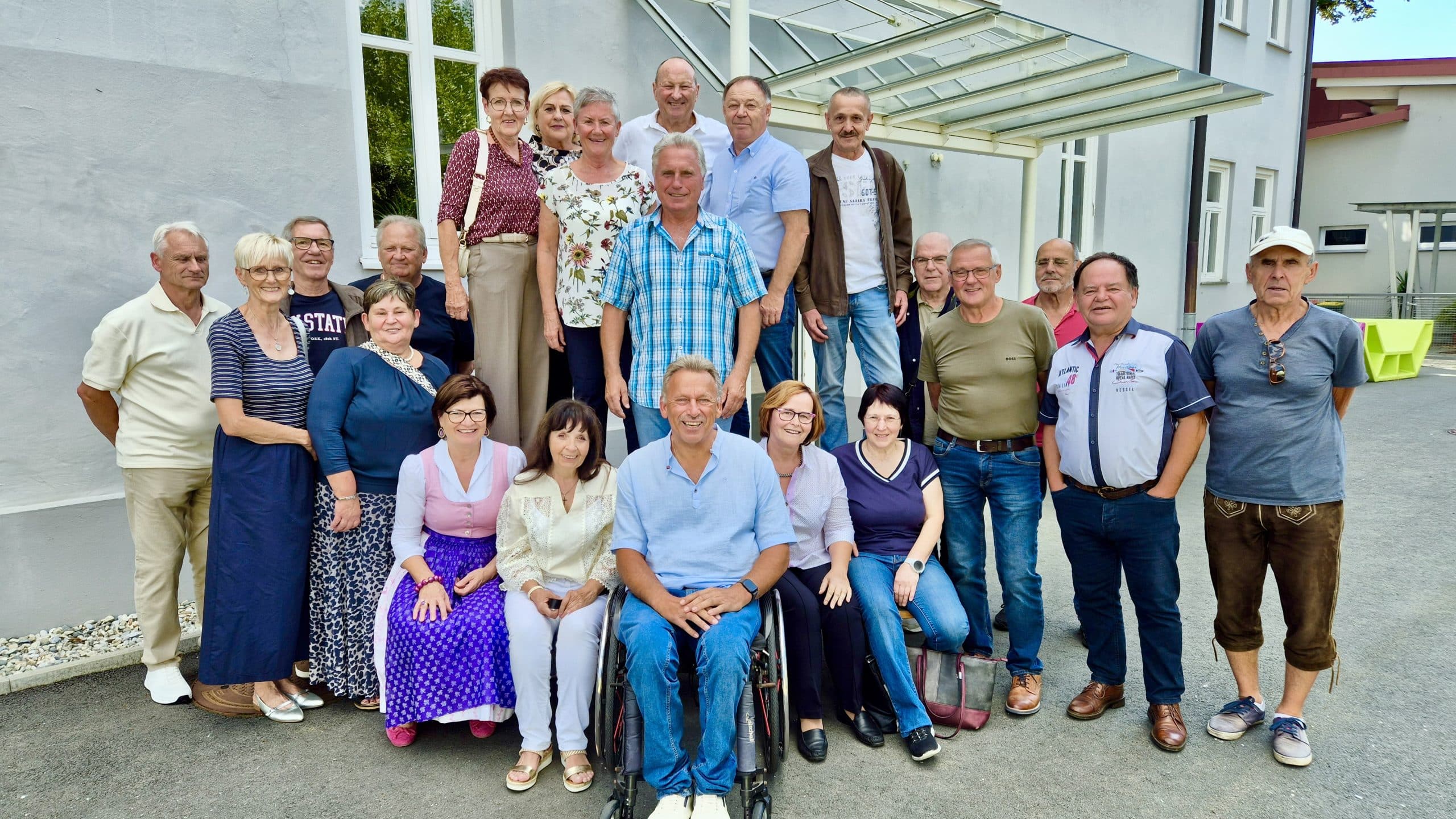 Eine Gruppe Erwachsener posiert gemeinsam im Freien vor einem Gebäude; einige stehen, andere sitzen, und ganz vorne in der Mitte sitzt ein Mann im Rollstuhl.