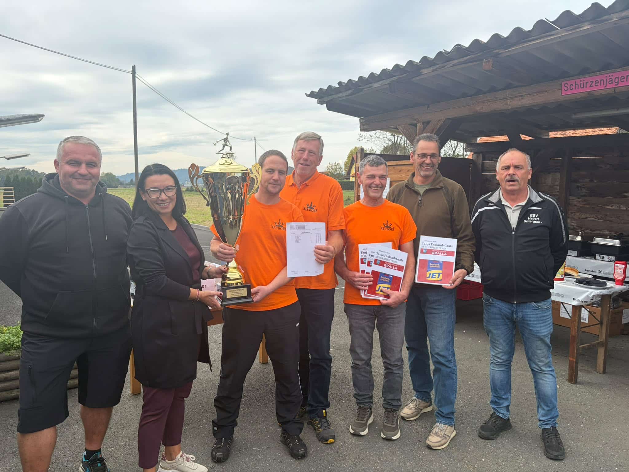 Sechs Personen stehen gemeinsam im Freien; ein Mann hält eine große goldene Trophäe in der Hand, die anderen halten Papiere und Urkunden in den Händen.