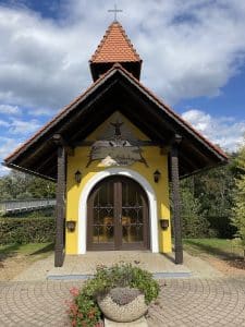 Eine gelbe Kapelle am Wegesrand mit Ziegeldach, gewölbten Flügeltüren und einem Wandgemälde des Heiligen Franz von Assisi über dem Eingang.