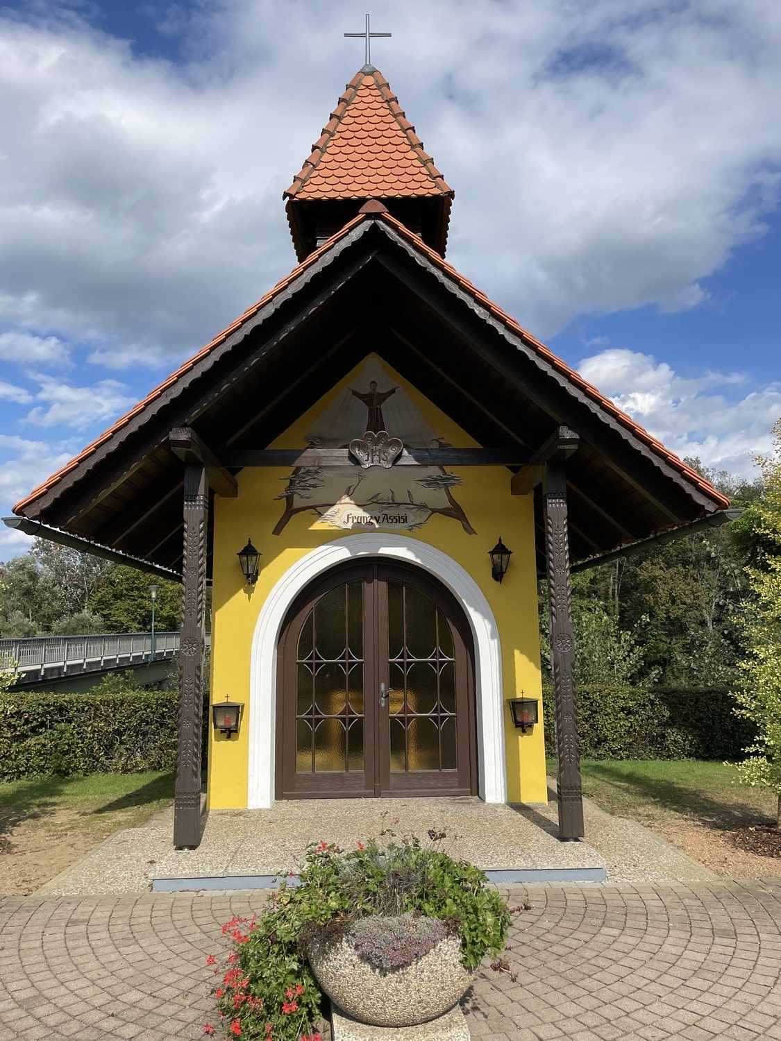 Eine gelbe Kapelle am Wegesrand mit Ziegeldach, gewölbten Flügeltüren und einem Wandgemälde des Heiligen Franz von Assisi über dem Eingang.