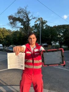 Ein Mitarbeiter des Österreichischen Roten Kreuzes in roter Uniform hält ein Papierformular und ein Tablet zur Dateneingabe in die Höhe.